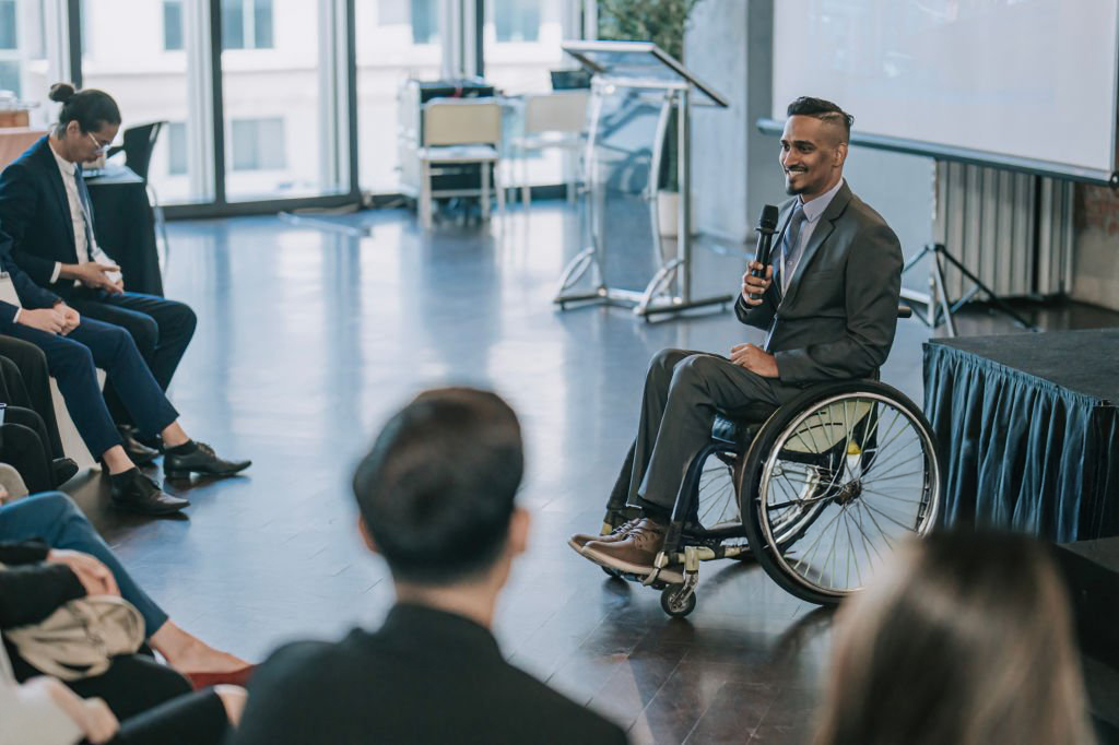 Businessman in a wheelchair giving a speech at a corporate event in a convention centre.