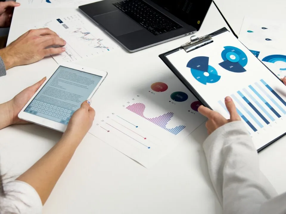 People collaborating at a table with a laptop, a tablet, and event marketing data charts on paper. A clipboard shows blue data visuals. The mood is focused.