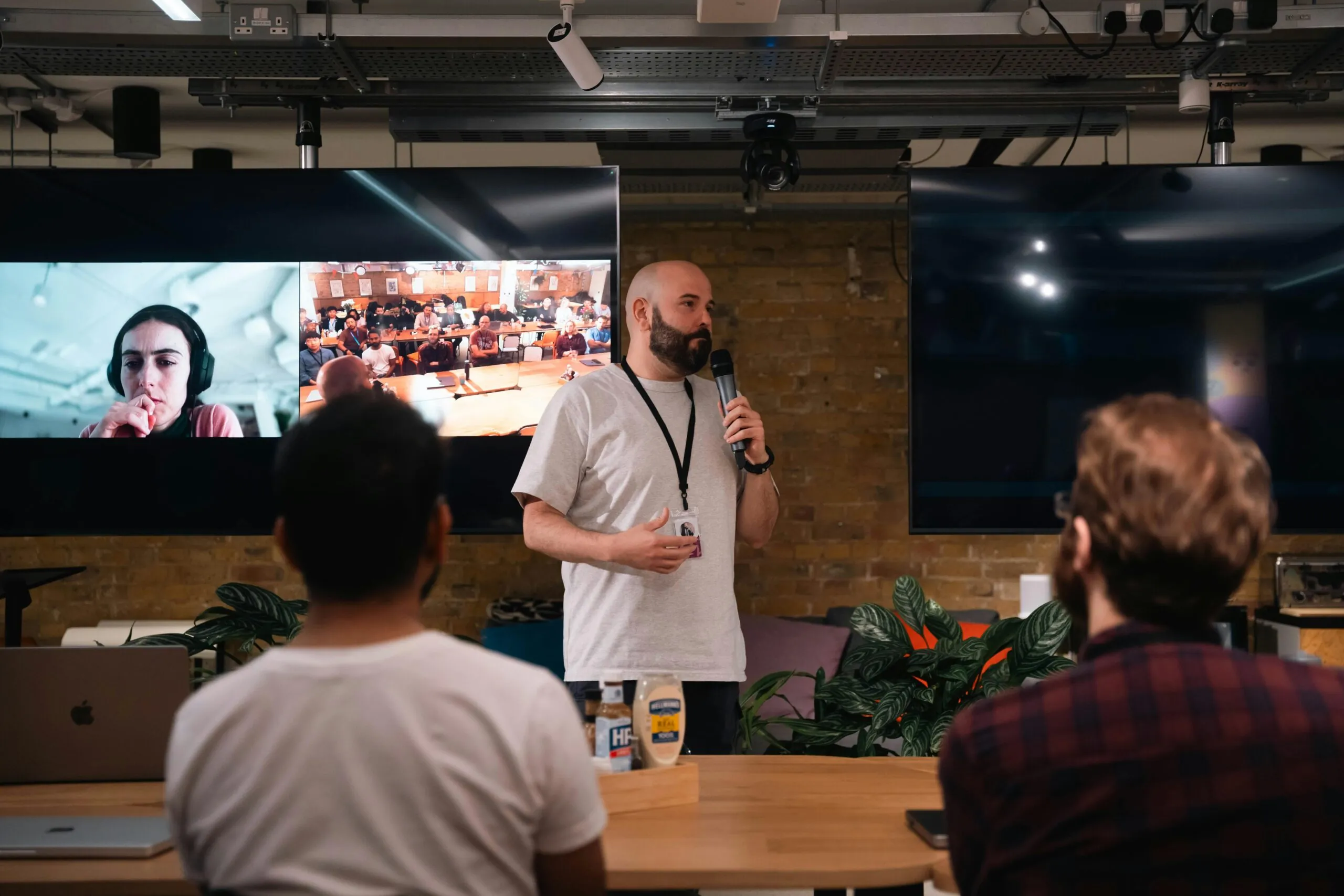 A speaker holding a microphone in front of an audience and digital screens showing hybrid engagement during an event marketing session.