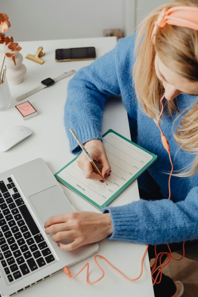 A woman in a blue sweater writes on a planner at a desk. She wears orange headphones, and her laptop is open beside her.