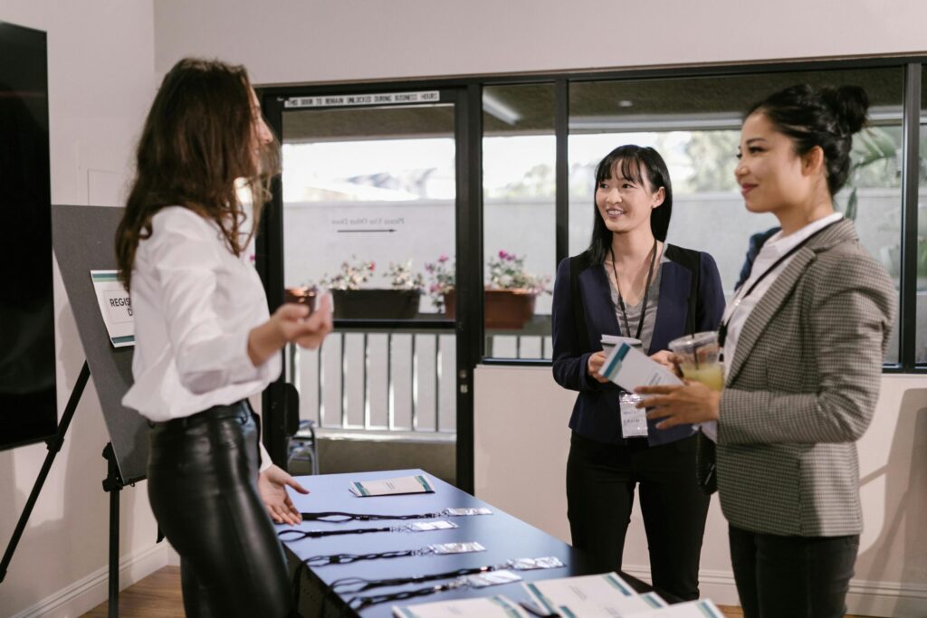 Three women at an event conference registration table. One event organiser, in a white blouse, assists two others in business attire, holding name tags and drinks, smiling.