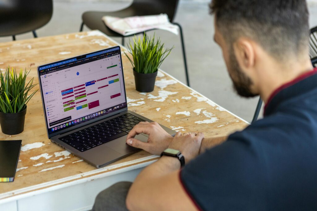 A man works on a laptop displaying a colorful event organiser calendar app, seated at a rustic table with small potted plants, conveying productivity and organization.