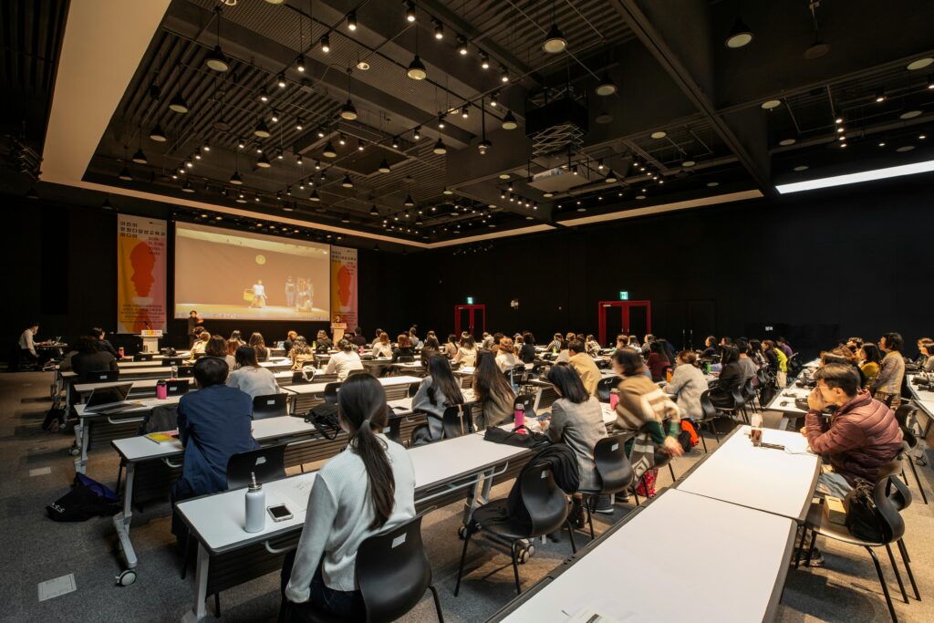 A large conference and seminar room filled with event attendees seated at rows of tables, facing a stage with a presentation screen.