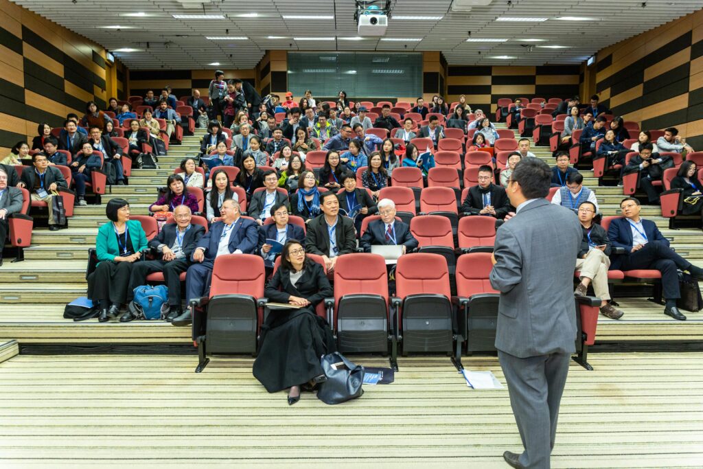 A speaker addresses a diverse audience seated in a corporate event venue hall. The crowd, engaged and attentive, fills the seats.