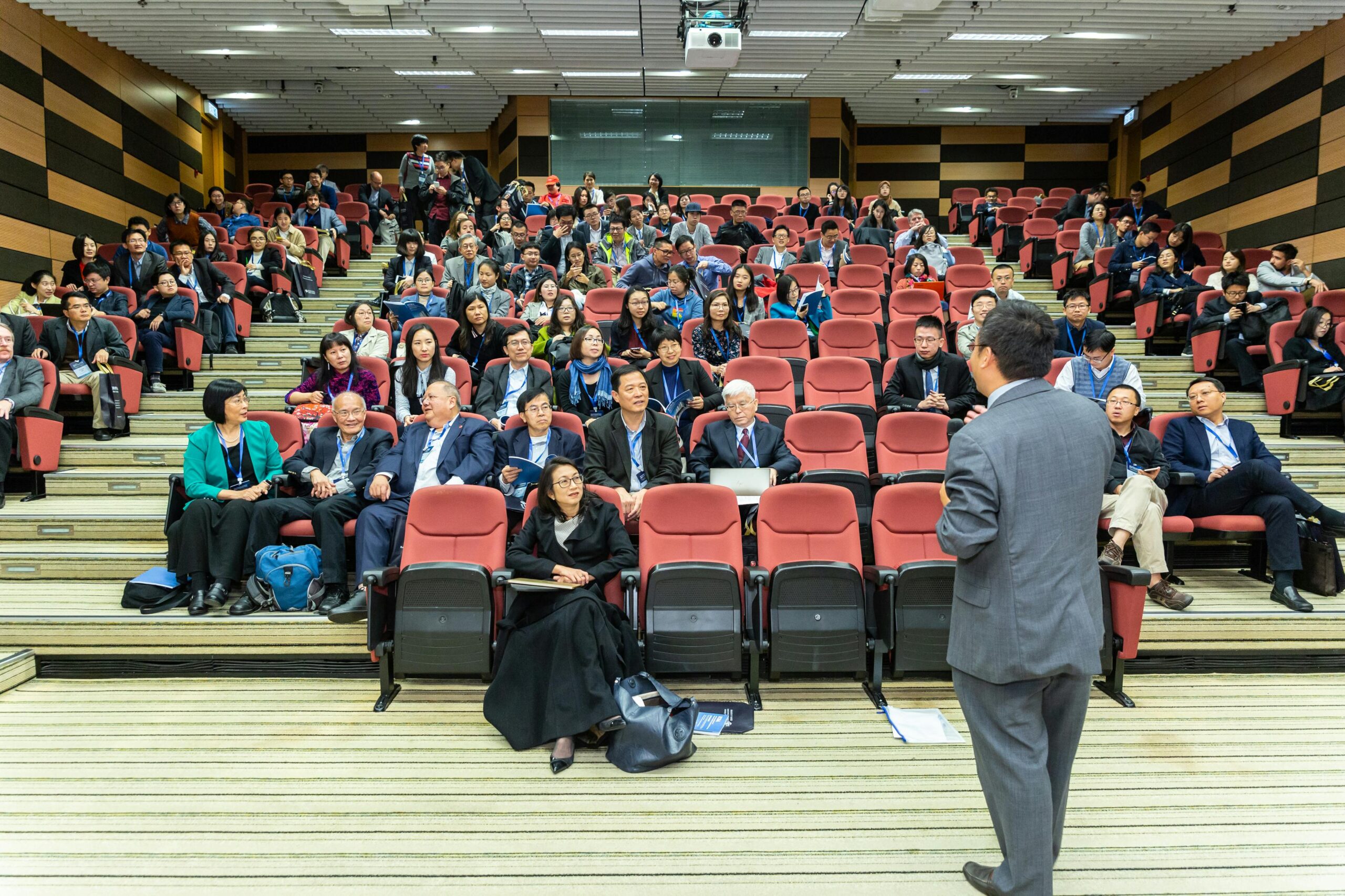 A speaker addresses a diverse audience seated in a corporate event venue hall. The crowd, engaged and attentive, fills the seats.