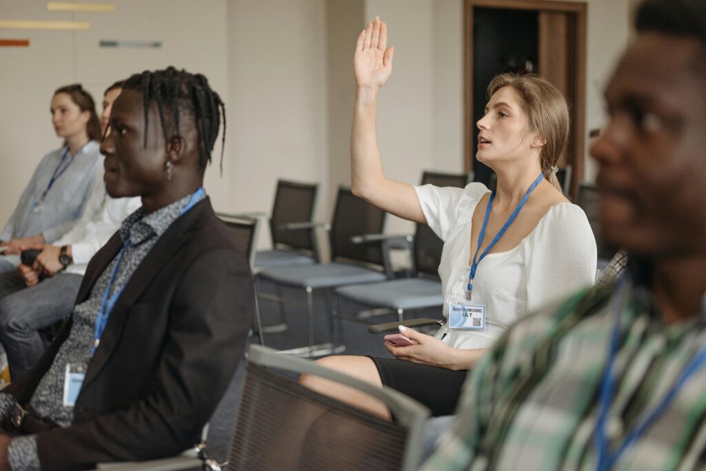 A woman in a white shirt raises her hand during a seminar or conference, conveying engagement.