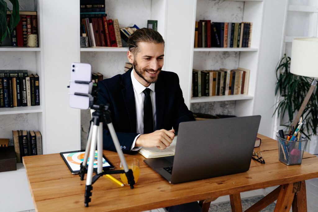 Smiling man in a suit sitting at a wooden desk with a laptop, phone on tripod, and tablet.