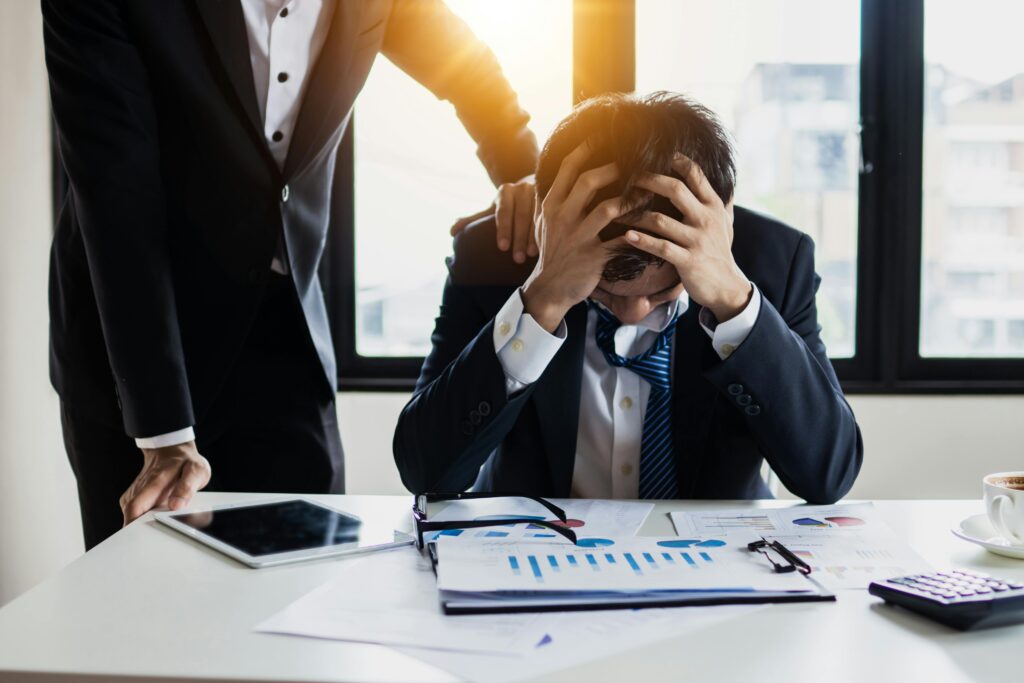 A stressed corporate event planner in a suit sits at a cluttered desk with graphs, head in hands. A colleague stands beside him, offering a reassuring touch.