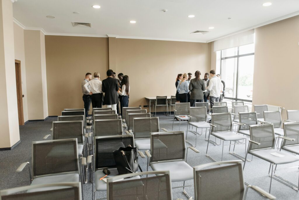 A group of people stands in a semi-circle talking in a networking event conference room with rows of empty chairs. The atmosphere appears casual and collaborative.