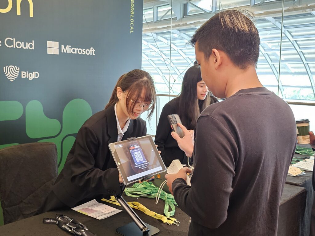 A woman helps a man scan an event registration system QR code on a tablet at a tech conference booth.