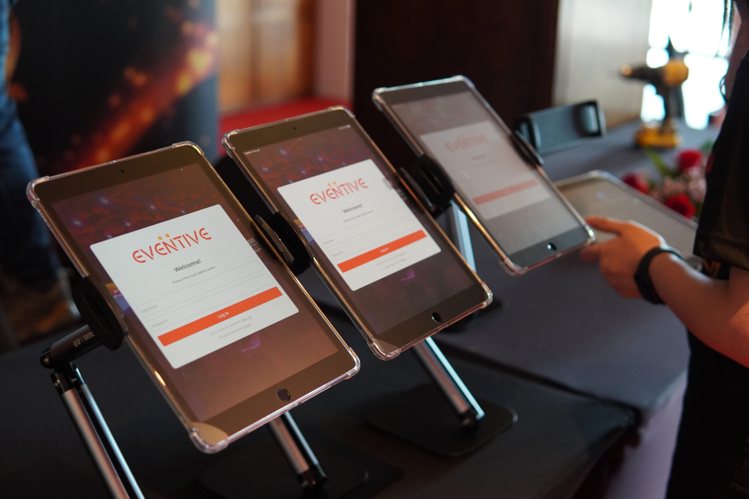 Three tablets on stands display a digital event registration system for "Eventive." A person interacts with the devices.