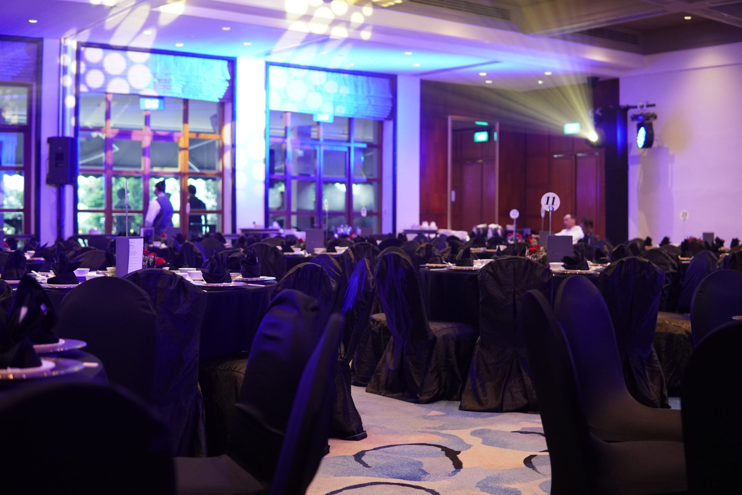 Elegant dinner and dance banquet hall with dim blue lighting, round tables draped in black cloths, and numbered table signs.