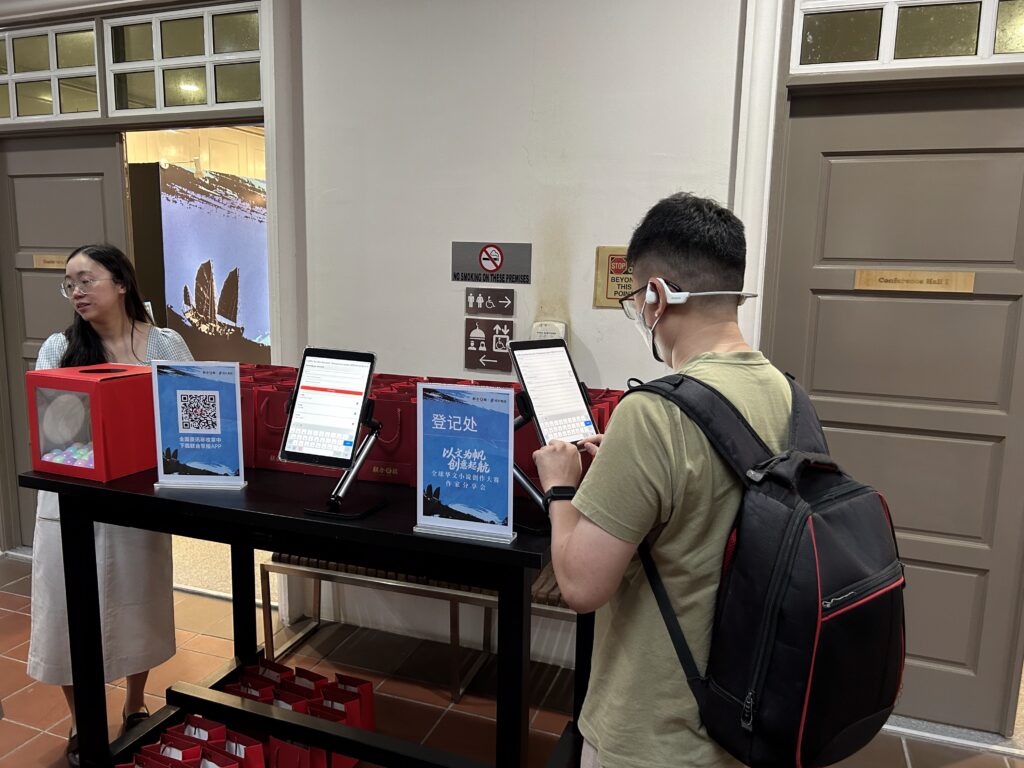 A man with a backpack fills in an event registration system form on a touchscreen device on a table in a public space.